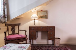 Een kamer met een houten bureau, een stoel met kussen, een lamp, een lijst met afbeelding en een kamerbreed tapijt met dessin.