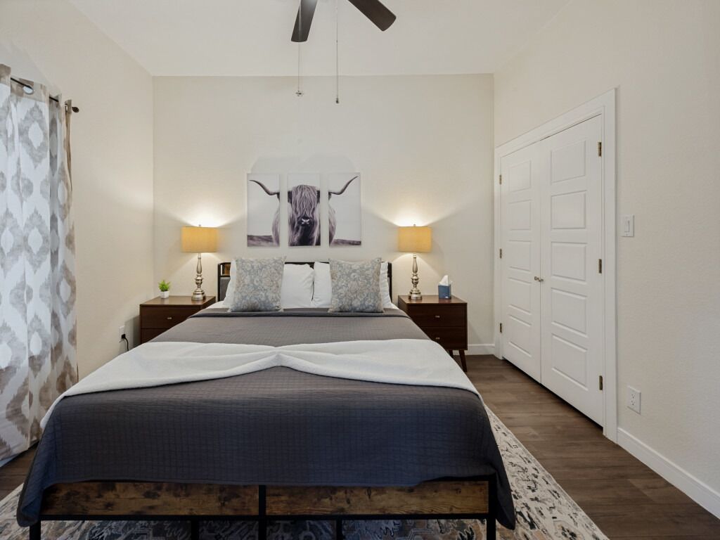 Une chambre à coucher comprenant un lit, deux tables de chevet, un ventilateur de plafond, une lampe fixée au mur, une porte et un tapis à motifs.