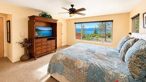 A bedroom with a bed, a TV on a wooden cabinet, a ceiling fan, and a window with a mountain view.
