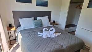 A neatly made bed with a quilted grey bedspread, a headboard, and a pair of white towels arranged in a swan shape on the bed. - Cube Exclusive 4 (Velsen-Zuid)