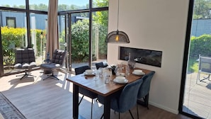 A modern dining area with a wooden table, blue chairs, and a large window offering a view of greenery and a building. - Cube Exclusive 4 (Velsen-Zuid)