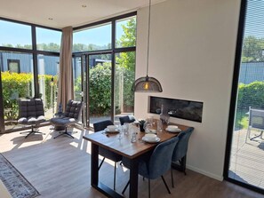 A modern dining area with a wooden table, blue chairs, and a large window offering a view of greenery and a building. - Cube Exclusive 4 (Velsen-Zuid)