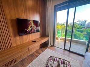 A modern living room with a flat-screen TV mounted on a wooden wall, a remote control on a wooden console, a patterned rug, and a large sliding glass door leading to a balcony with a view of greenery and buildings. - Marrakech-style villa with pool, hammam, and sauna (Amizmiz)