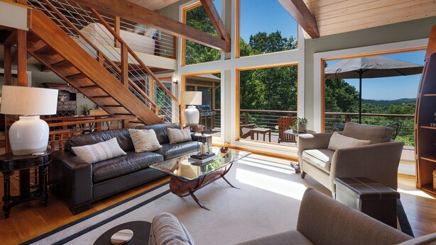 Amplia sala de estar con escalera, sofá de cuero, mesa de centro de vidrio y ventanas grandes que ofrecen vistas al exterior.