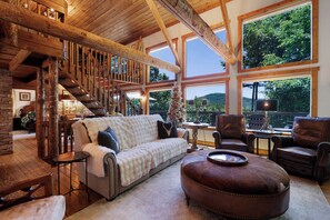 A spacious living area with a wooden staircase, a sofa, leather chairs, and large windows offering a view of the outdoors. - Chalet Lumiere - Big Canoe, GA (Jasper)