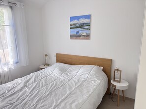 Ein Schlafzimmer mit einem Bett, einem Nachttisch, einer Lampe, einem Bild von einer Strandansicht und einem Fenster mit Vorhängen.