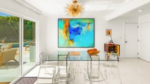 Et moderne spiseområde med et glasbord, transparente stole, et stort abstrakt maleri og en markant lampe.
