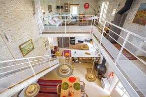 Un interior a varios niveles con cocina, comedor y sala de estar, que presenta una pared de piedra, barandillas blancas y un dormitorio en la entreplanta.