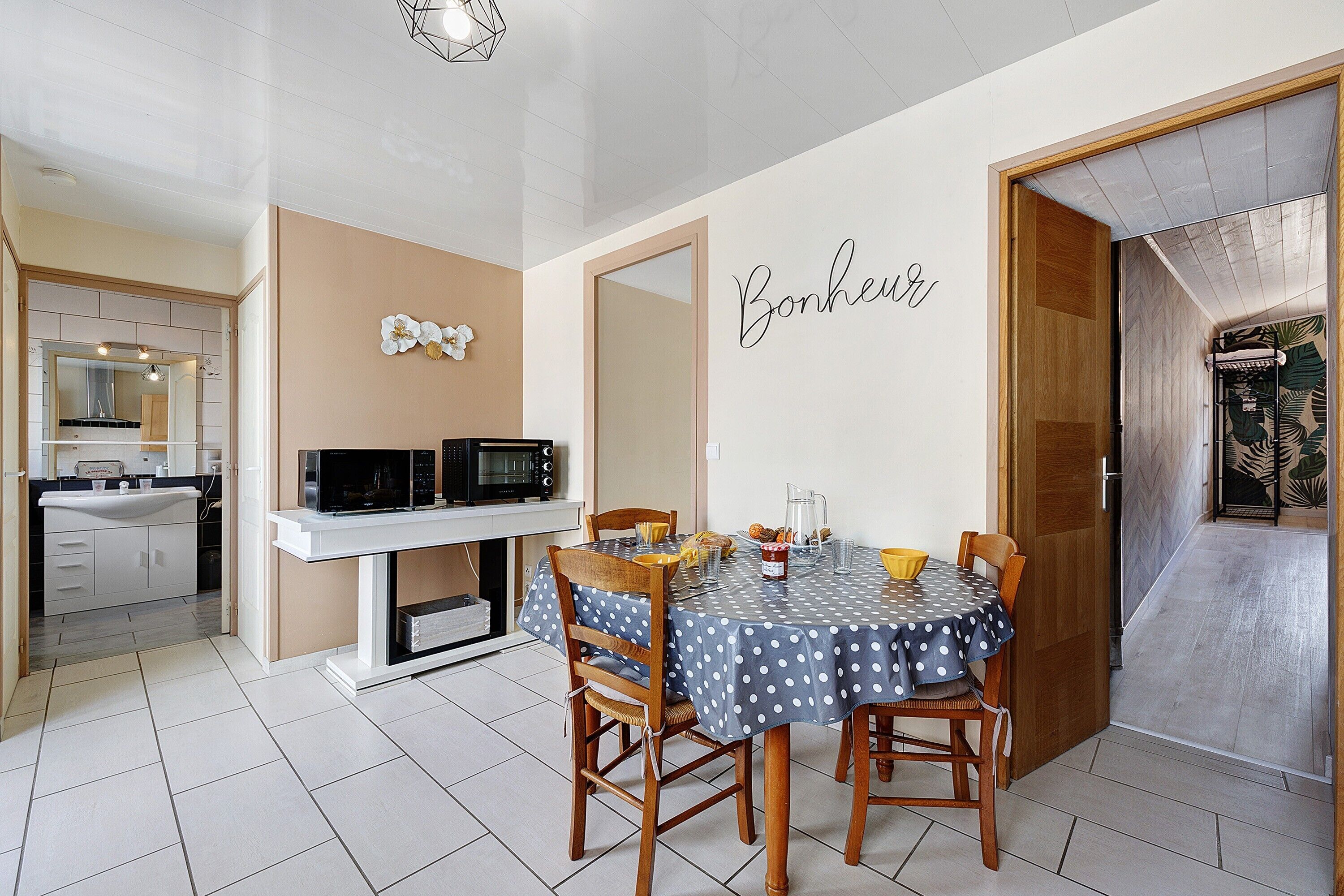 Un área de comedor con una mesa preparada para cuatro, una cocina al fondo y una puerta que da a un baño con ducha.