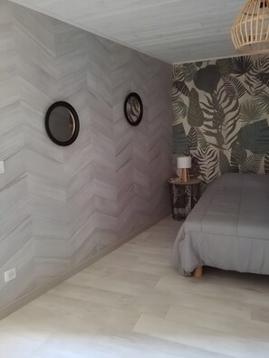 Et soveværelse med en chevron-mønstret væg, en seng med gråt sengetøj, et rundt spejl, et lille natbord med en lampe, og en væg med et tropisk blad-motiv.