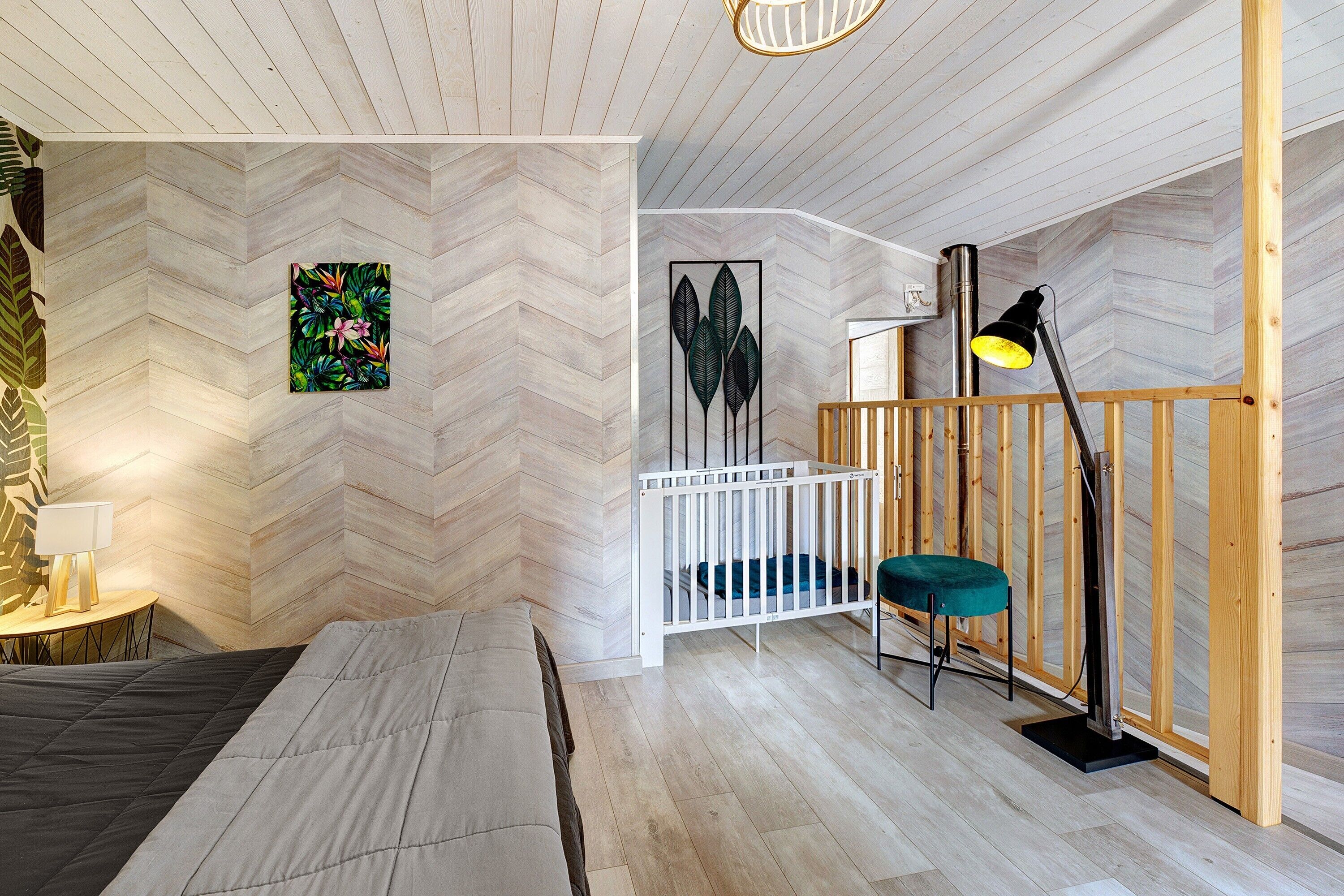 Quarto com parede em padrão herringbone, uma cama, um berço, uma escrivaninha com luminária e uma planta pendente.