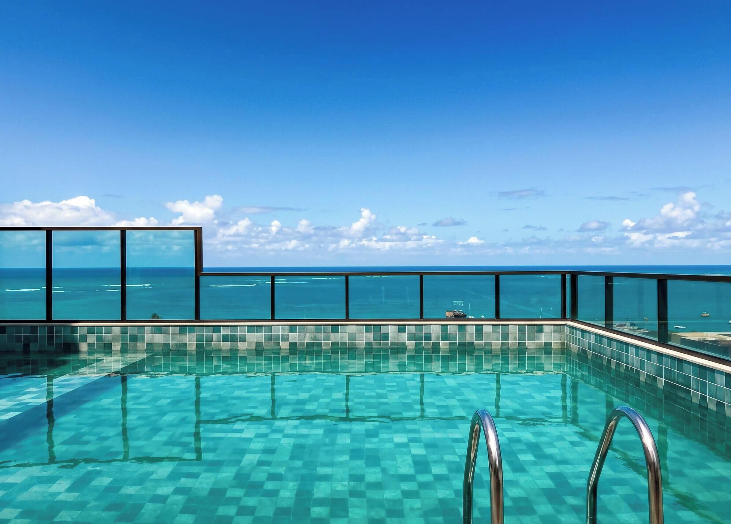 A clear blue sky, a swimming pool with checkered tiles, and a view of the ocean.