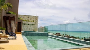 Une piscine à débordement située sur le toit, offrant une vue sur l’océan, un espace détente avec des fauteuils et un bâtiment moderne orné d’un mur en mosaïque.