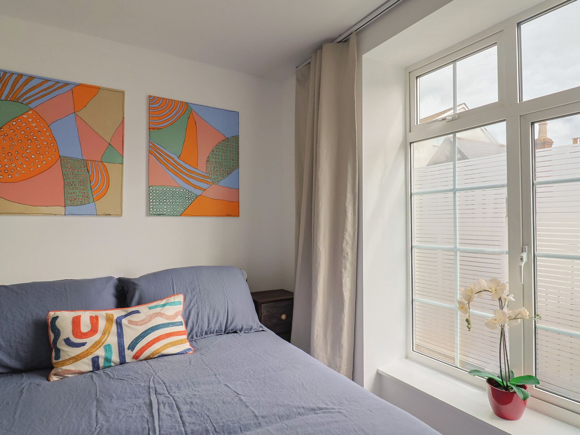 Um quarto com uma cama, almofadas, uma janela com cortinas e duas pinturas abstratas na parede.