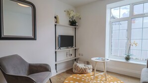 Una sala de estar con un sillón gris, un tapete redondo con lunares blancos, un televisor en un estante, una mesita y una ventana con persianas.