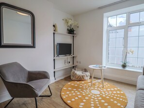 Una sala de estar con un sillón gris, un tapete redondo con lunares blancos, un televisor en un estante, una mesita y una ventana con persianas.