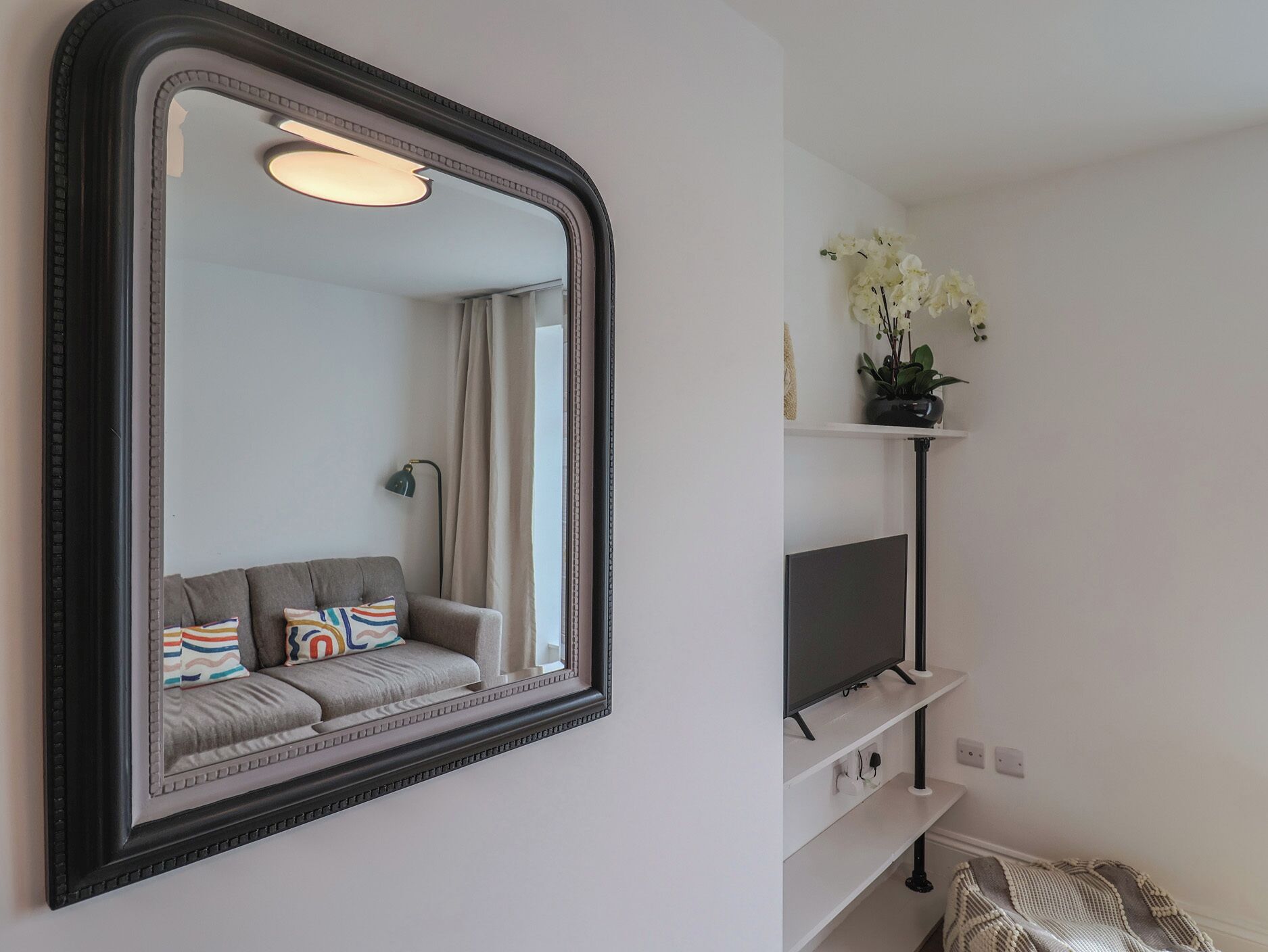 A room with a large mirror, a grey sofa with colorful pillows, a television on a white shelf, and a potted plant with white flowers.