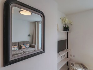 Una habitación con un espejo grande, un sofá gris con almohadones de colores, un televisor sobre un estante blanco y una planta en maceta con flores blancas.