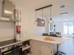 A modern kitchen with a built-in stove, overhead ventilation, pendant lighting, and a dining area with a table and chairs. - Flat 1 Harley House (Sidmouth)