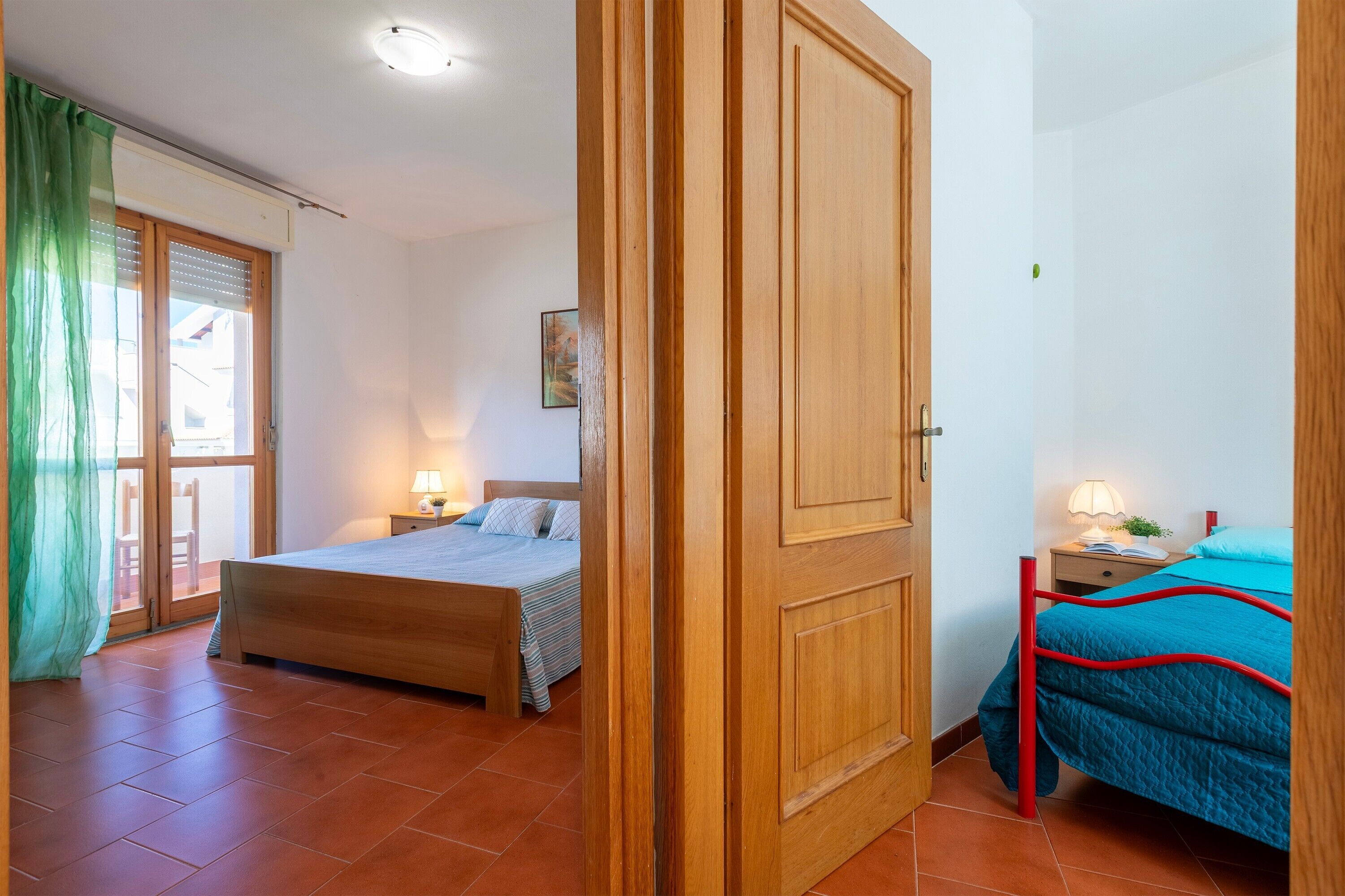 Ein Schlafzimmer mit einem Bett, einem Nachttisch, einer Lampe, einem Fenster mit Vorhängen und einer Tür.