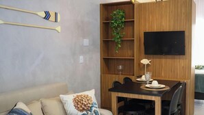 Une pièce comprenant une table à manger en bois, des chaises, un téléviseur fixé au mur, une bibliothèque agrémentée d’une plante et un canapé orné de coussins décoratifs.