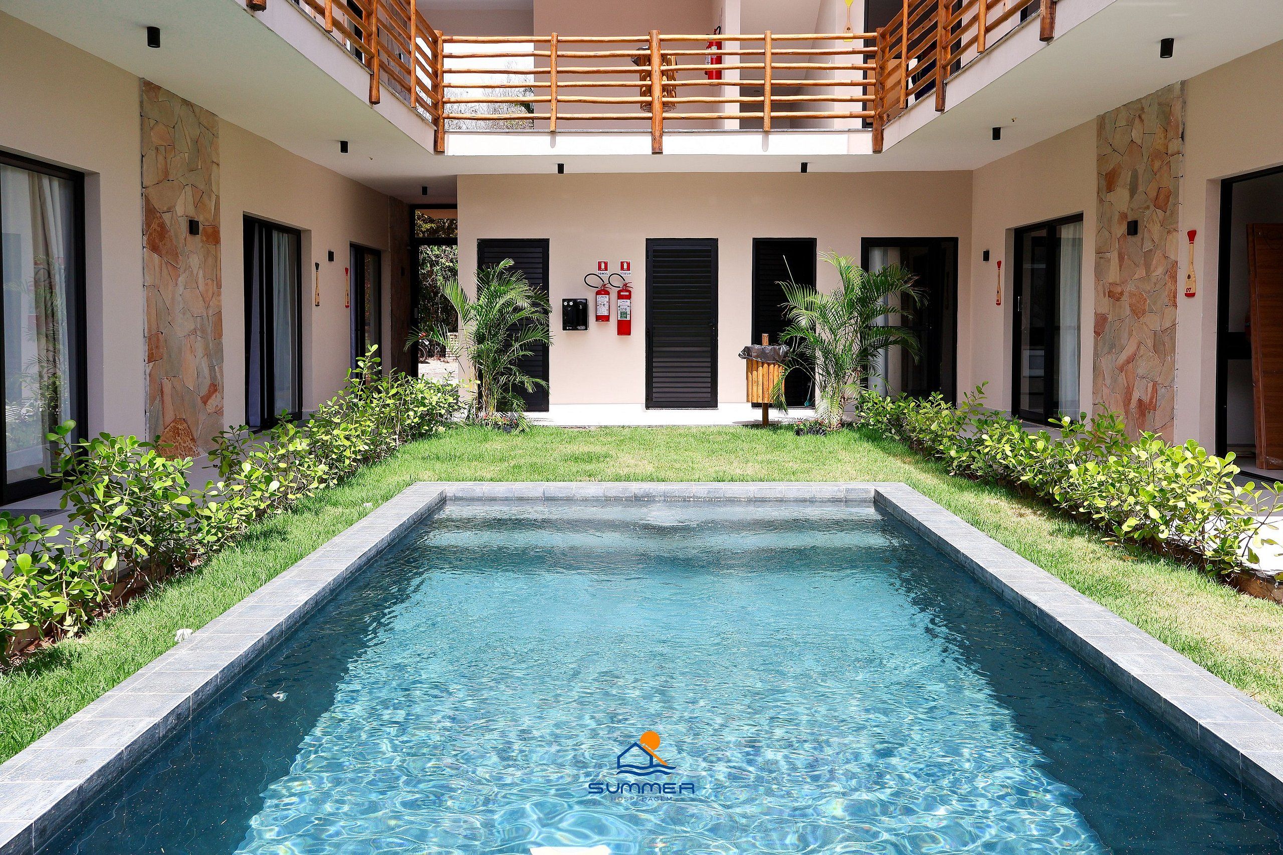 A swimming pool with a logo, a covered outdoor area with a wooden railing, and a stone wall with doors.