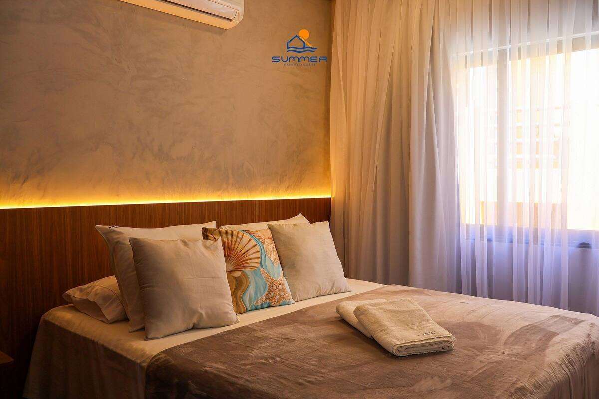 Habitación de hotel con cama, cabecera, ventana con cortinas traslúcidas y un logotipo en la pared.