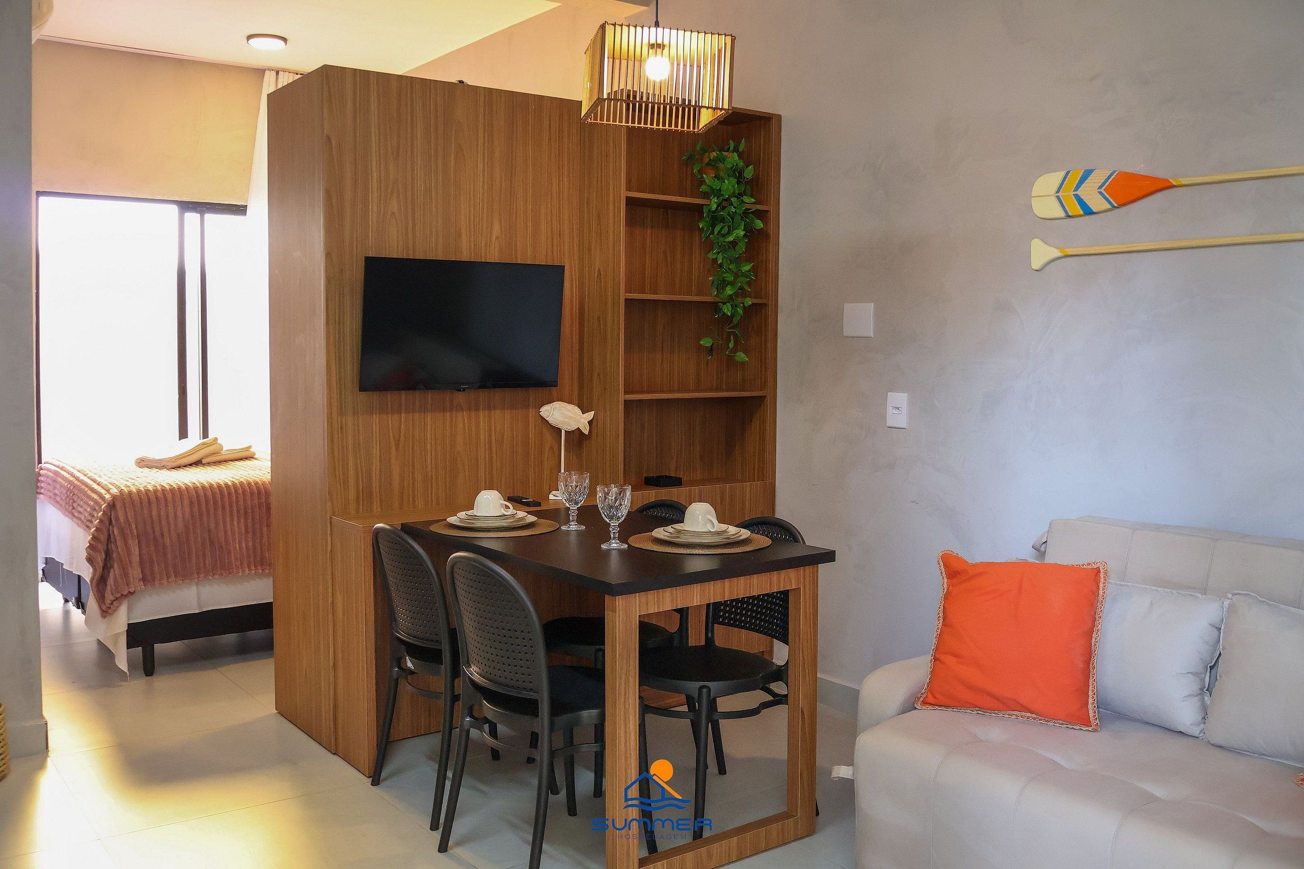 A compact living space with a dining area, a TV mounted on the wall, a plant, and a couch with an orange pillow.