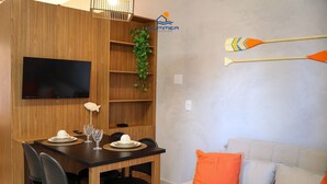A modern room with a wooden dining table, chairs, a flat-screen TV, a sofa with orange pillows, and a wall-mounted paddleboard. - Condominio Petit (TIBAU DO SUL)