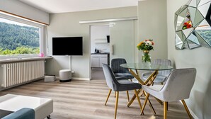Un salon moderne avec une table à manger en verre, des chaises grises et blanches, un téléviseur à écran plat et une vue sur une forêt, visible à travers une grande fenêtre.