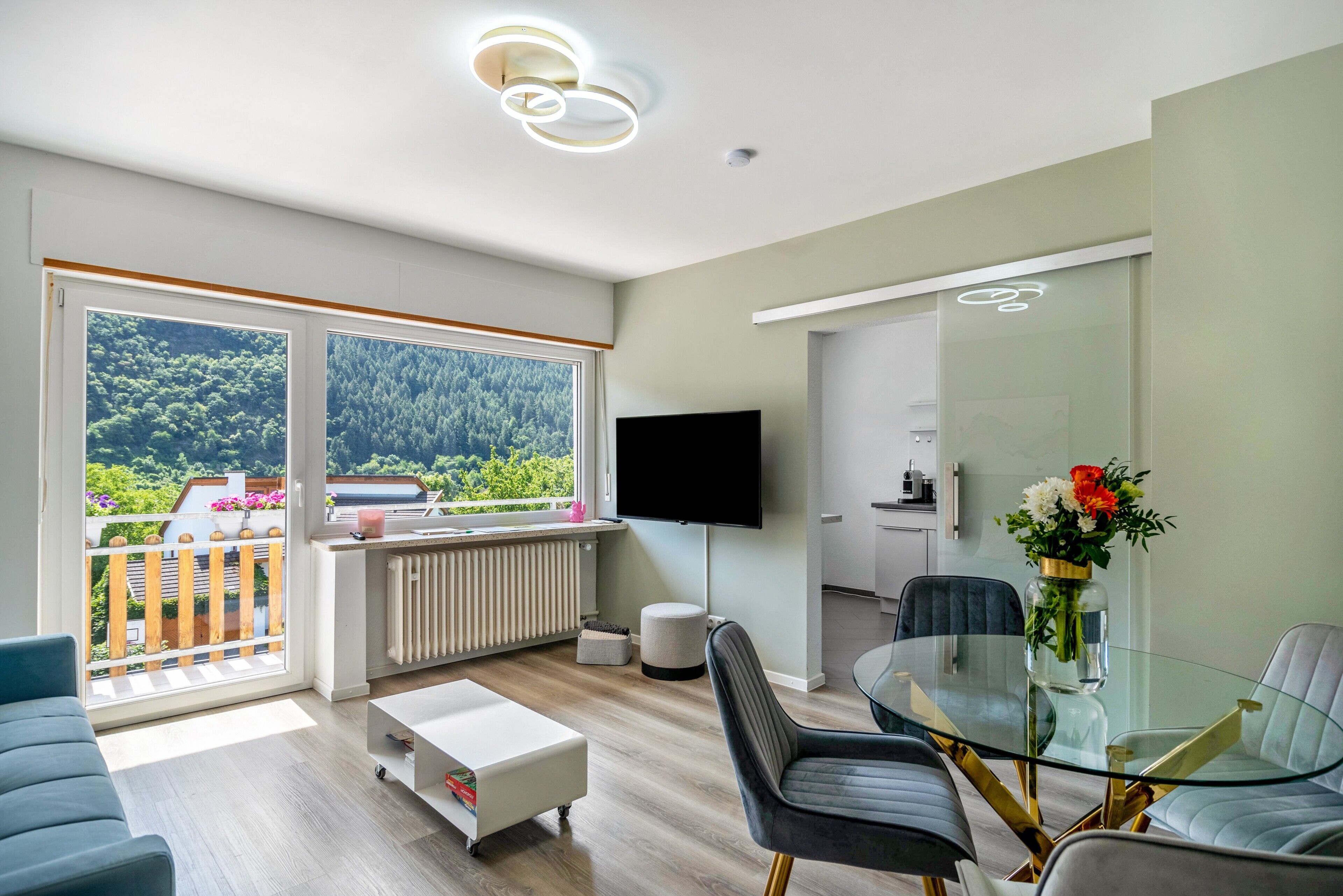 A modern living room with a large window, a flat-screen TV, a glass dining table, and a sofa.