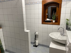 Ein Badezimmer mit weißen Fliesen, einem spiegelnden Holzrahmen, einem Waschbecken und einem Toilettenpapierhalter.