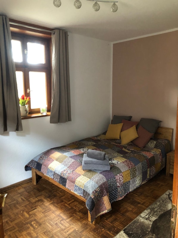 Ein Schlafzimmer mit Holzboden, einem Bett mit einer gesteppten Decke, einem Fenster mit Vorhängen und einem kleinen Tisch mit Blumen in einer Vase.