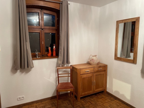 A corner room with a wooden cabinet, a chair, a window with curtains, and a mirror on the wall.