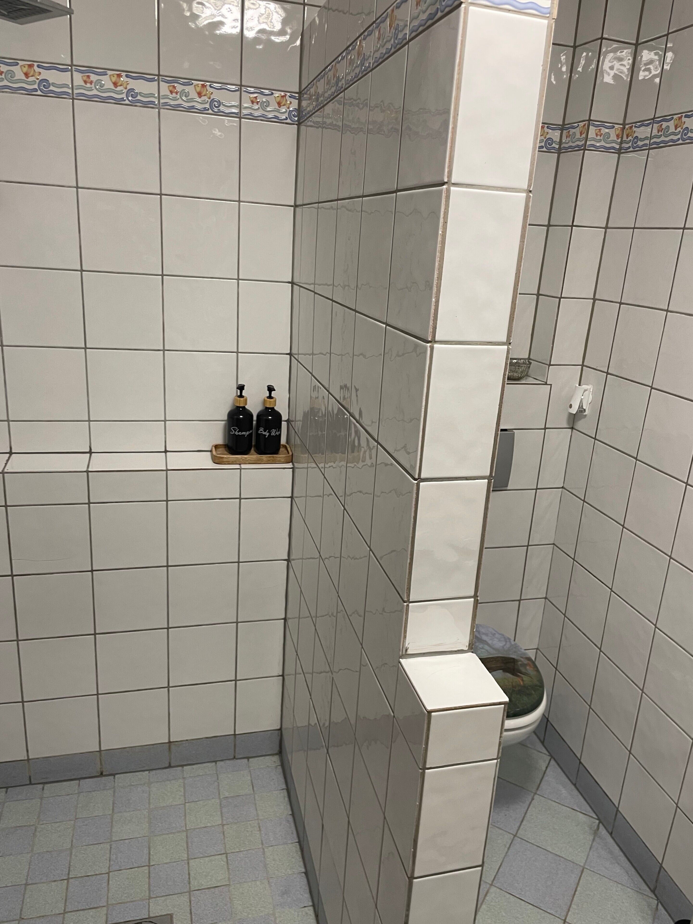 Une salle de bain avec des carreaux blancs, des toilettes et deux bouteilles posées sur une étagère.