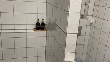 Une salle de bain avec des carreaux blancs, des toilettes et deux bouteilles posées sur une étagère.