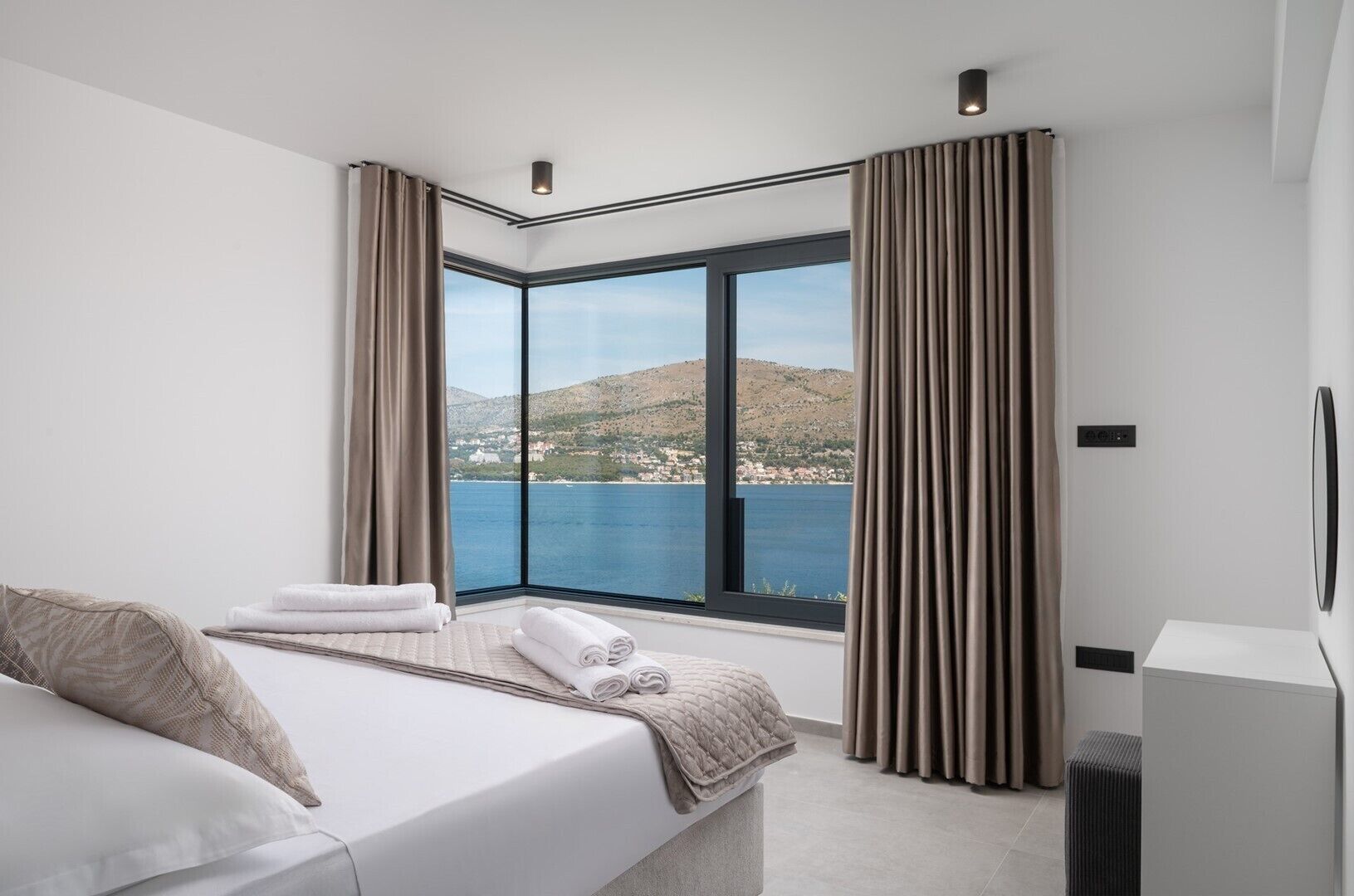 Ein modernes Hotelzimmer mit großem Fenster, einem Bett, einem Nachttisch und Blick auf das Meer und die Hügel.