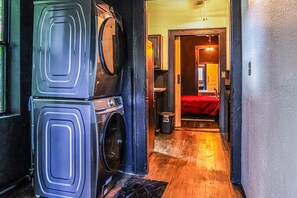 A laundry area with stacked washers and dryers, a wooden floor, and a bed with a red cover in the adjacent room. - Frenchmen - New Orleans (New Orleans)