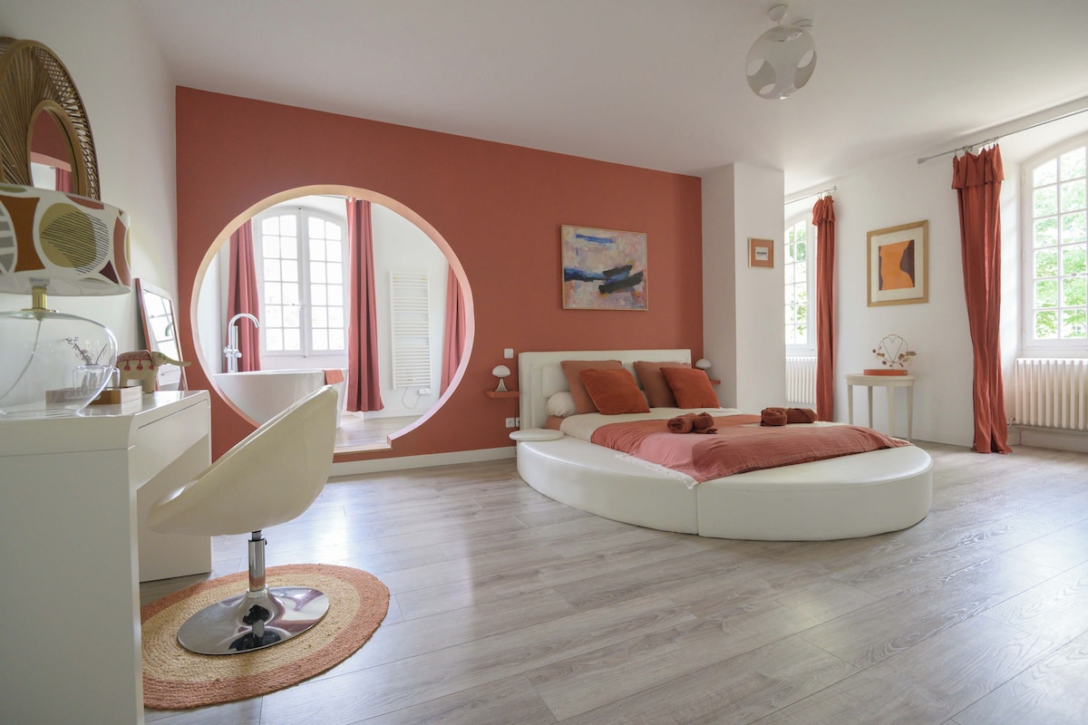 Een slaapkamer met een rond bed, een wit bureau, een spiegel en een raam met gordijnen.