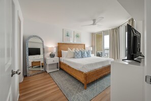 Ein Schlafzimmer mit Bett, Nachttischen, Fernseher, Deckenventilator und einem großen Fenster mit Vorhängen.