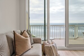 A room with a large window overlooking the ocean, a grey sofa with cushions, and a wicker basket on the floor. - Beautifully Designed Oceanfront Villas Close to Everything (Folly Beach)