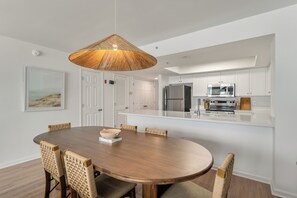 A modern kitchen with white cabinetry, a large island, and a dining area with a round wooden table and woven chairs. - Beautifully Designed Oceanfront Villas Close to Everything (Folly Beach)