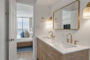 Une salle de bain avec un double vasque, des robinetteries dorées, un grand miroir et une vue sur la pièce.
