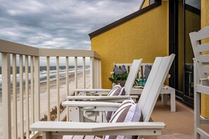 En balkon med hvide stole og et bord, med udsigt over en strand og bølger.