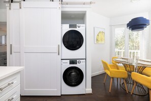 A modern kitchen with a built-in washer and dryer, a dining area with yellow chairs, and a large window with blinds. - Charming Ocean View Condo in Wild Dunes (Isle Of Palms)