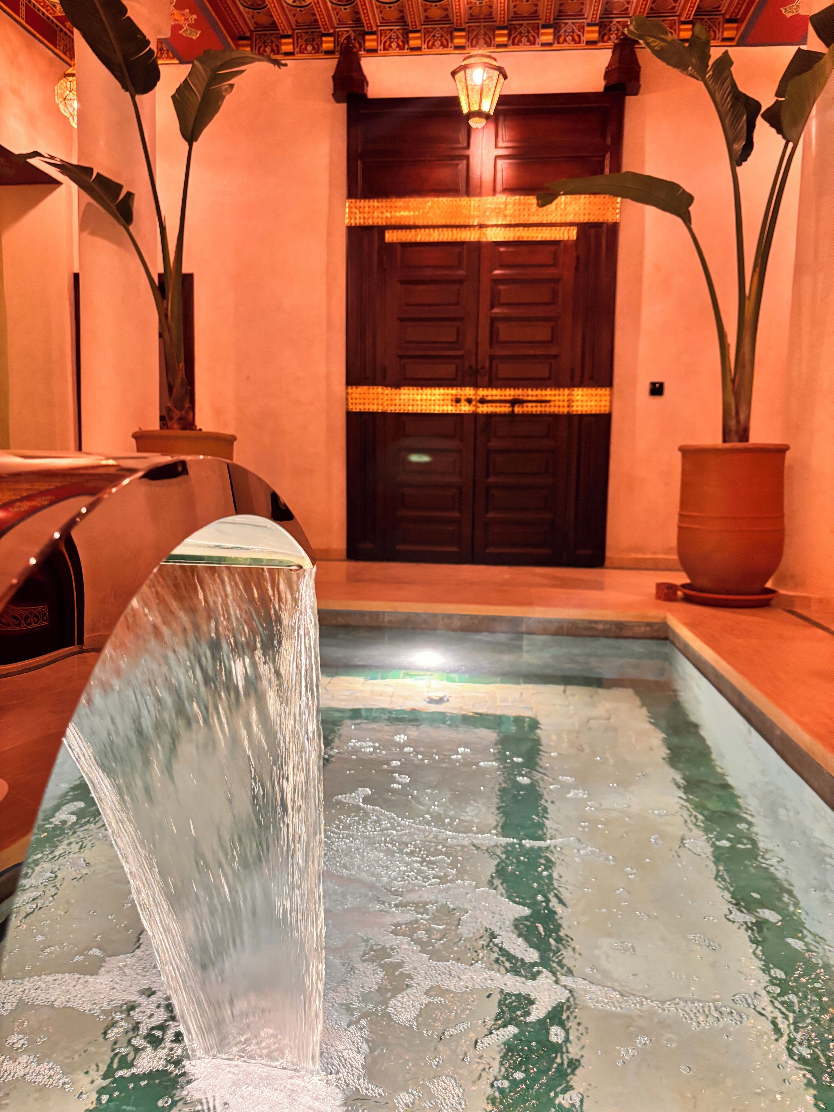 Una fuente con una cascada que desemboca en una piscina, rodeada de plantas en macetas y un banco de madera.