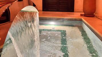 Una fuente con una cascada que desemboca en una piscina, rodeada de plantas en macetas y un banco de madera.