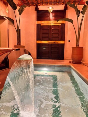 A water feature with a cascading waterfall in a pool, surrounded by potted plants and a wooden bench.