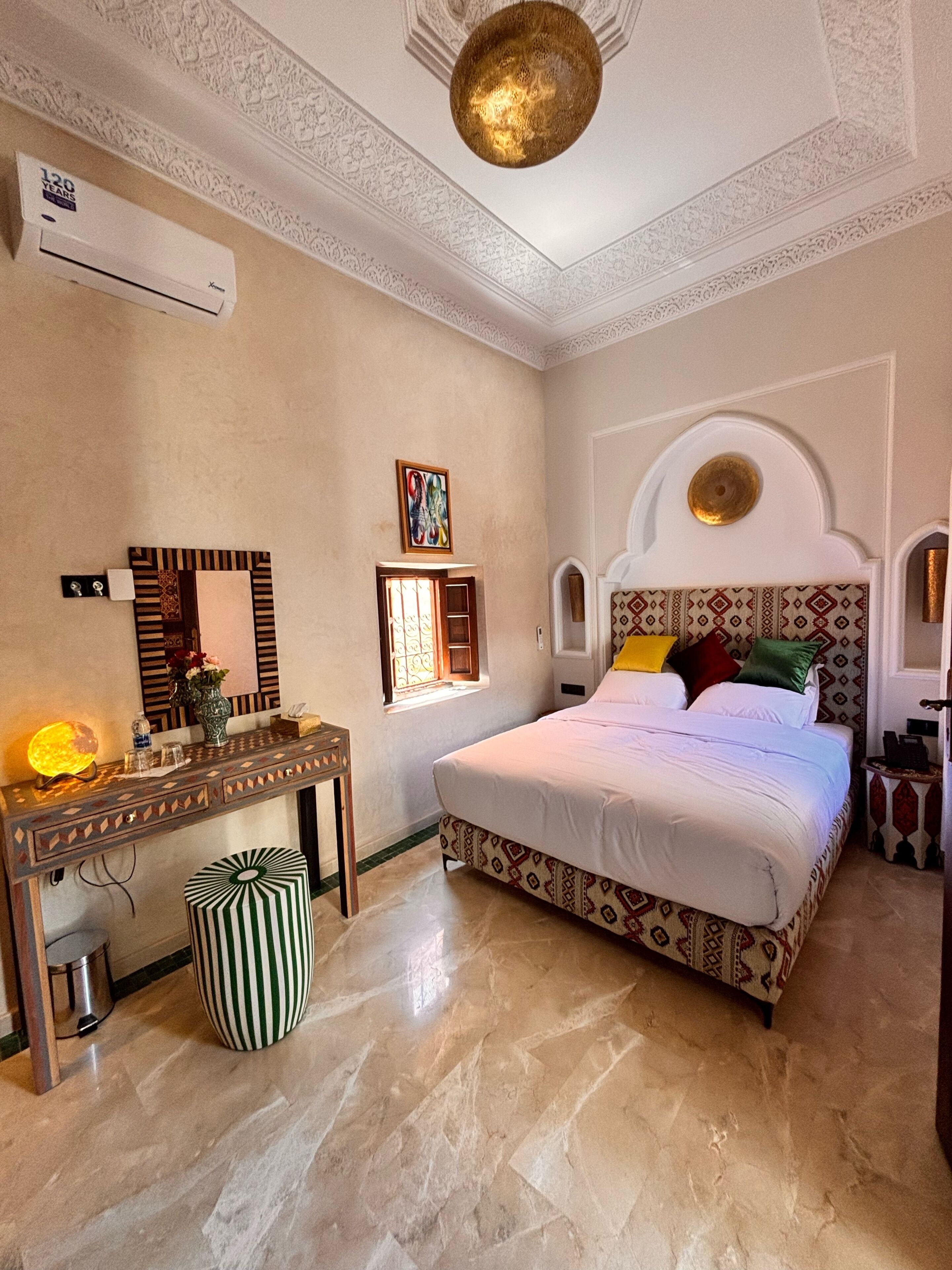 Habitación de hotel con cama, tocador con espejo, aire acondicionado de pared y cabecero estampado.