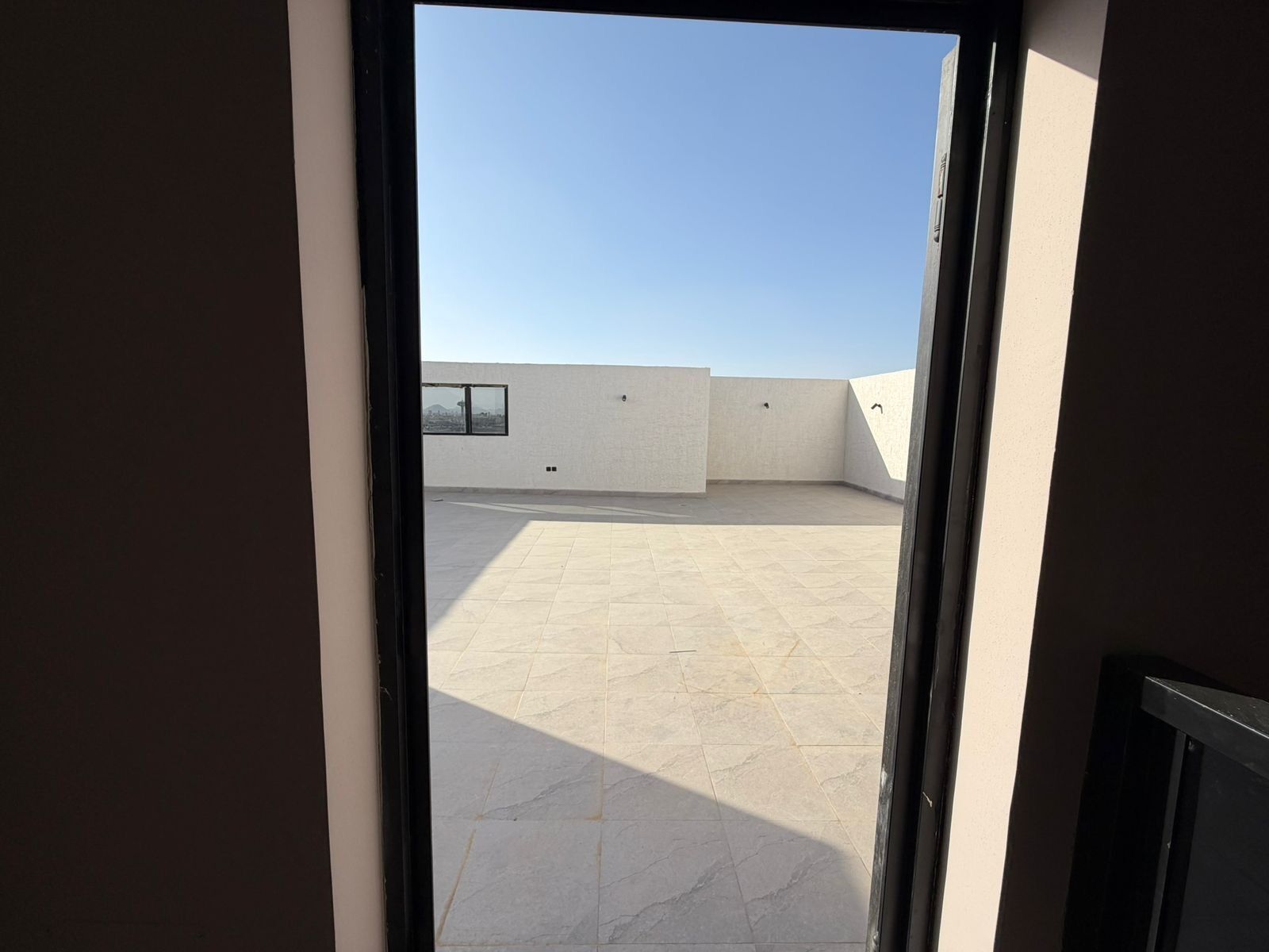 A view through a doorway onto a rooftop terrace with a clear sky, a window, and a wall.